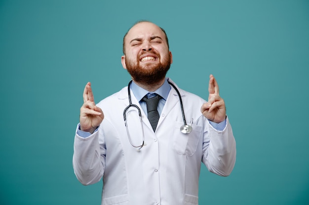 Jeune médecin de sexe masculin plein d'espoir portant un manteau médical et un stéthoscope autour du cou faisant un vœu avec les yeux fermés et les doigts croisés isolés sur fond bleu