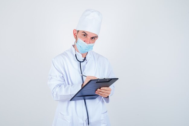 Jeune médecin de sexe masculin prenant des notes sur le presse-papiers en uniforme blanc, masque et à la vue sensible, de face.