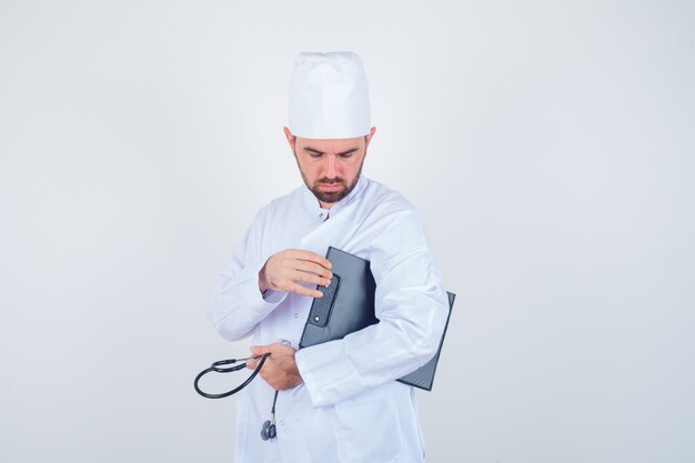 Jeune médecin de sexe masculin tenant le presse-papiers et le stéthoscope en uniforme blanc et à la vue réfléchie, de face.