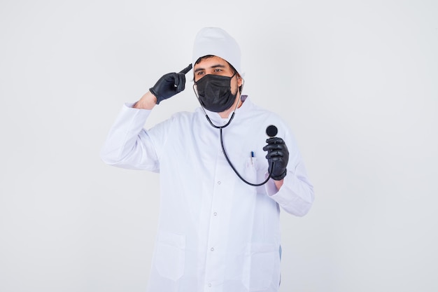 Jeune médecin de sexe masculin en uniforme blanc pointant la tête tout en tenant un stéthoscope et en ayant l'air confiant, vue de face.