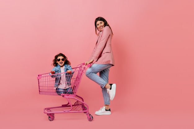 Jeune mère raffinée avec enfant posant après le shopping. Vue pleine longueur de la famille avec chariot de magasin.