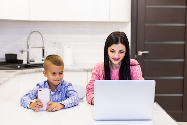 Jeune mère travaillant sur ordinateur portable à la maison, tandis que son enfant s'ennuie à jouer sur le téléphone