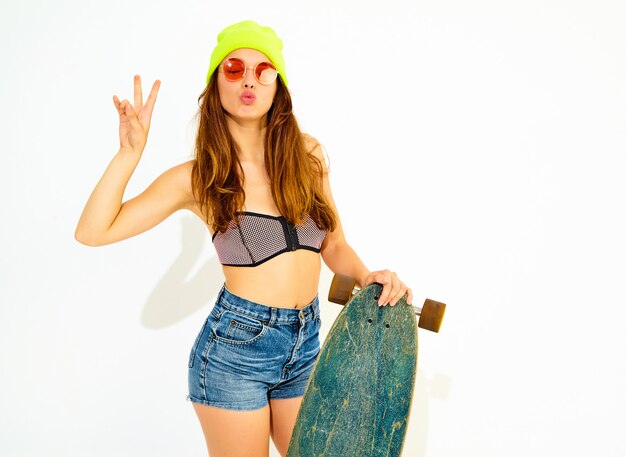 Jeune modèle de femme souriante élégante en vêtements de bain d'été décontractés et bonnet jaune posant avec bureau longboard, montrant le signe de la paix. Isolé sur blanc