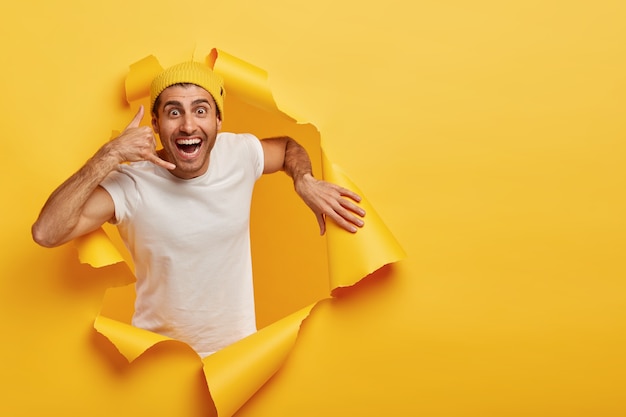 Jeune modèle masculin attrayant positif fait un geste d'appel, porte un chapeau jaune et un t-shirt blanc décontracté