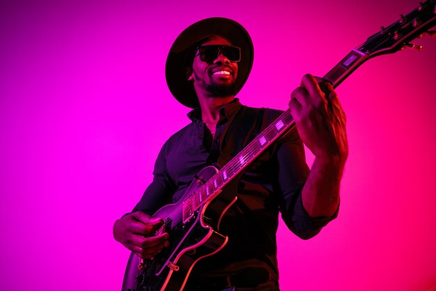 Jeune musicien afro-américain jouant de la guitare comme une rockstar sur un mur dégradé violet-rose à la lumière du néon