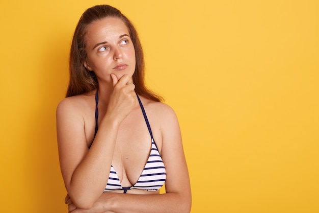 Jeune nageuse européenne isolée sur un mur jaune, a l'air pensif, regardant l'espace de copie avec une expression faciale réfléchie, robes maillot de bain rayé.