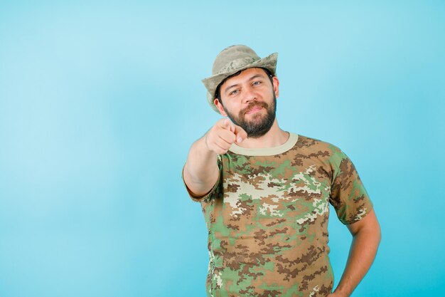 Le jeune officier pointe la caméra avec l'index et met l'autre main sur la taille sur fond bleu