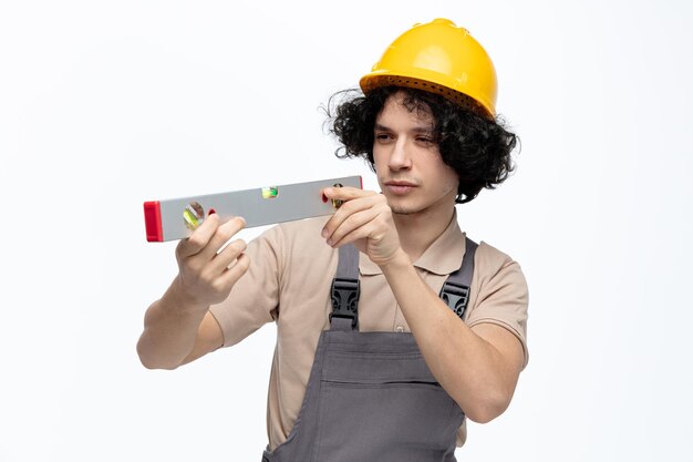 Jeune ouvrier du bâtiment concentré portant un uniforme et un casque de sécurité tenant et regardant au niveau à bulle isolé sur fond blanc