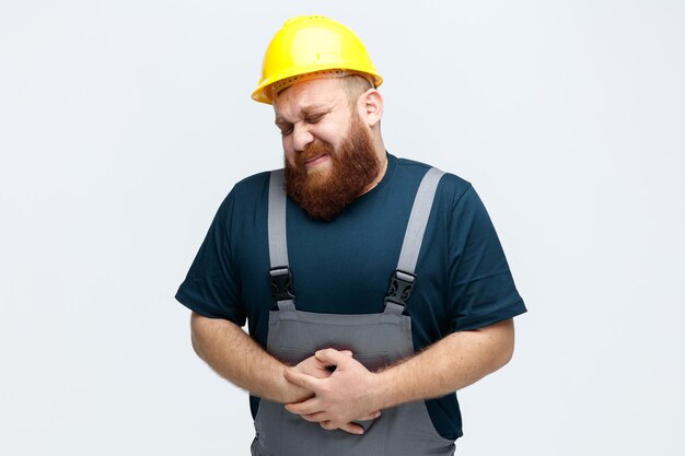 Jeune ouvrier du bâtiment endolori portant un casque de sécurité et un uniforme gardant les mains sur le ventre avec les yeux fermés isolé sur fond blanc