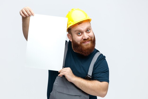 Jeune ouvrier du bâtiment inspiré portant un casque de sécurité et un uniforme regardant la caméra montrant du papier à la caméra isolé sur fond blanc