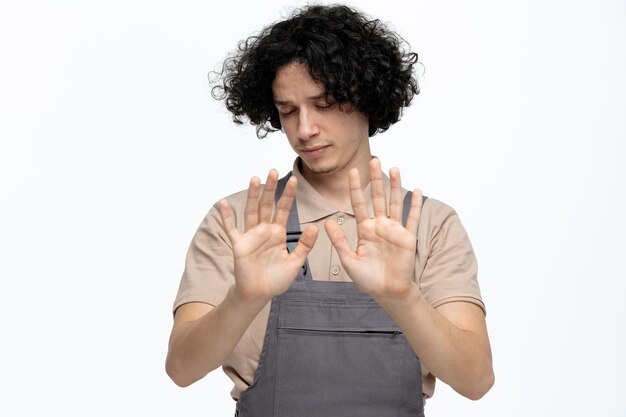 Jeune ouvrier du bâtiment mécontent portant l'uniforme faisant un geste d'arrêt avec les yeux fermés isolé sur fond blanc