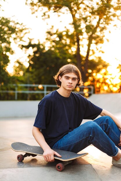 Jeune patineur en T-shirt noir et jeans regardant pensivement à huis clos avec planche à roulettes passant du temps au skate park moderne