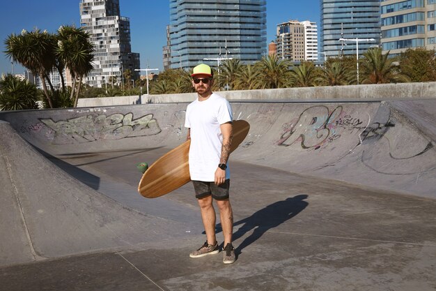 Jeune patineur tatoué attrayant en casquette de camionneur se dresse dans un t-shitrt blanc vierge sans étiquette avec son longboard en bois à la main au centre du skatepark, paysage urbain derrière