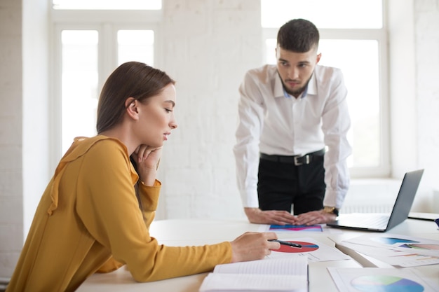 Jeune Patron Masculin En Chemise Blanche S'appuyant Sur La Table Tout En Regardant Pensivement L'employé Jolie Fille En Blouse Travaillant Avec Des Diagrammes Dans Un Bureau Moderne