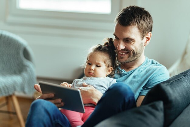 Jeune père heureux utilisant une tablette numérique avec sa petite fille tout en se relaxant dans le salon.