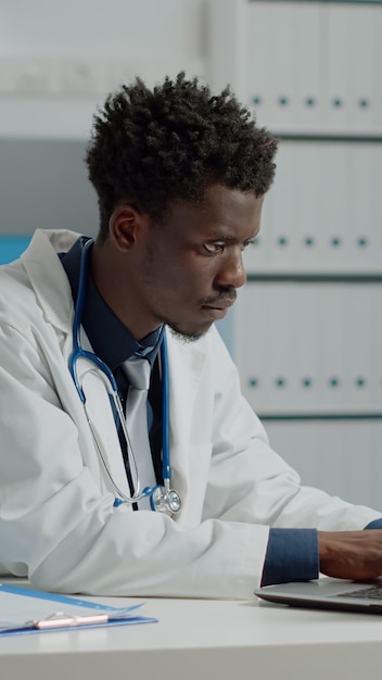 Jeune personne travaillant comme médecin dans un cabinet médical avec un ordinateur portable et des fichiers de documents. Medic portant une blouse blanche et un stéthoscope assis au bureau à l'aide de la technologie et des outils dans la chambre d'hôpital