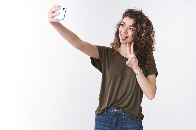 Jeune petite amie géorgienne amusante et mignonne aux cheveux bouclés prenant selfie envoyer son petit ami via l'application Internet étendre le bras vers le haut tenant le smartphone montrer la victoire de la langue ou le geste de paix, fond blanc