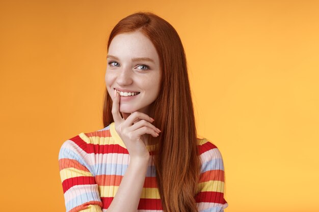 Une jeune petite amie rousse curieuse et sournoise de 20 ans a une excellente idée en souriant avec un sourire narquois aux lèvres tactiles délicates, une caméra mystérieusement en train de regarder la caméra a des plans pour préparer une surprise intéressante, un fond orange debout.