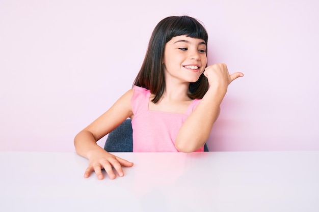 Photo gratuite jeune petite fille avec bang portant des vêtements décontractés assis sur la table souriant avec un visage heureux regardant et pointant vers le côté avec le pouce vers le haut