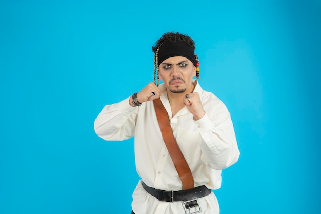 Un jeune pirate en colère serre ses pieds et regarde la caméra. Jeune homme debout fond bleu. Photo de haute qualité