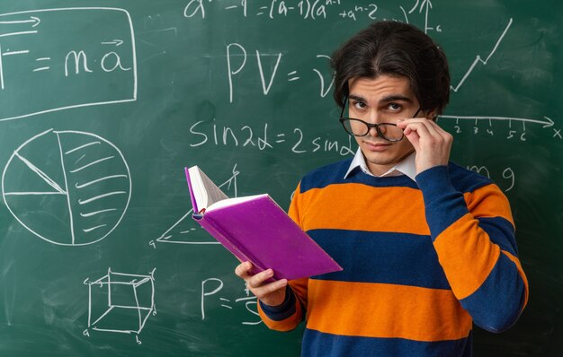 Jeune professeur de géométrie douteux portant des lunettes debout devant un tableau en classe regardant à l'avant tenant un livre ouvert en saisissant des lunettes