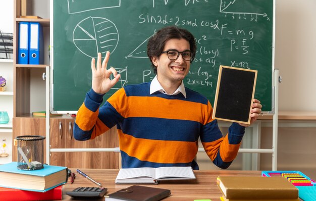Jeune professeur de géométrie joyeux portant des lunettes assis au bureau avec des fournitures scolaires en classe montrant un mini tableau noir regardant à l'avant faisant signe ok