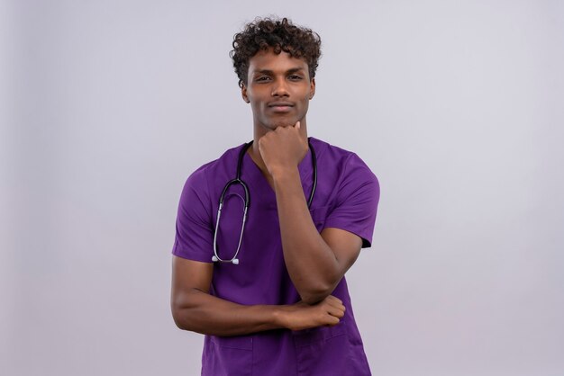 Un jeune sérieux beau médecin à la peau sombre avec des cheveux bouclés portant l'uniforme violet avec stéthoscope en gardant la main sur le menton