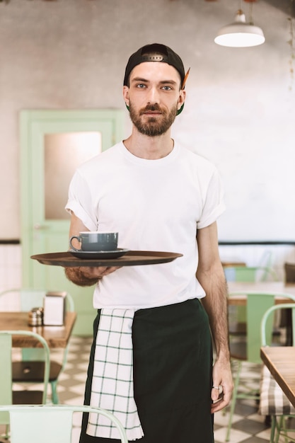 Photo gratuite jeune serveur cool en casquette noire et tablier debout et tenant un plateau avec une tasse de café tout en regardant rêveusement à huis clos au café
