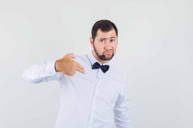 Jeune serveur faisant signe de pistolet à doigt pointé sur lui-même en chemise blanche et l'air confiant. vue de face.