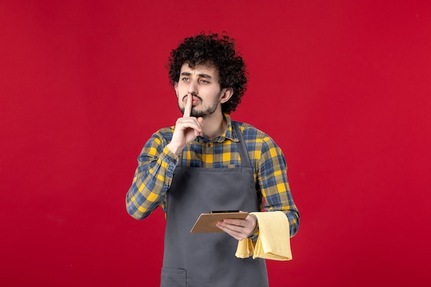 Jeune serveur masculin sérieux avec les cheveux bouclés tenant la serviette prenant l'ordre faisant un geste de silence sur fond rouge isolé