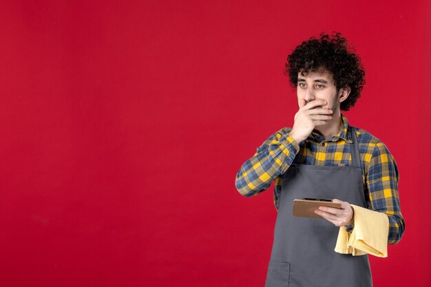 Jeune serveur masculin surpris avec des cheveux bouclés tenant une serviette prenant l'ordre sur fond rouge isolé