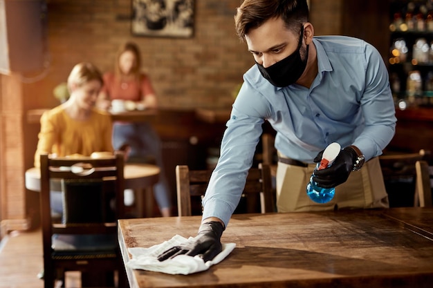 Jeune serveur portant un masque protecteur tout en nettoyant les tables tout en travaillant dans un café