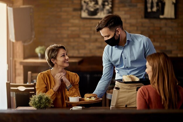 Jeune serveur portant un masque protecteur tout en servant de la nourriture à ses invités dans un restaurant