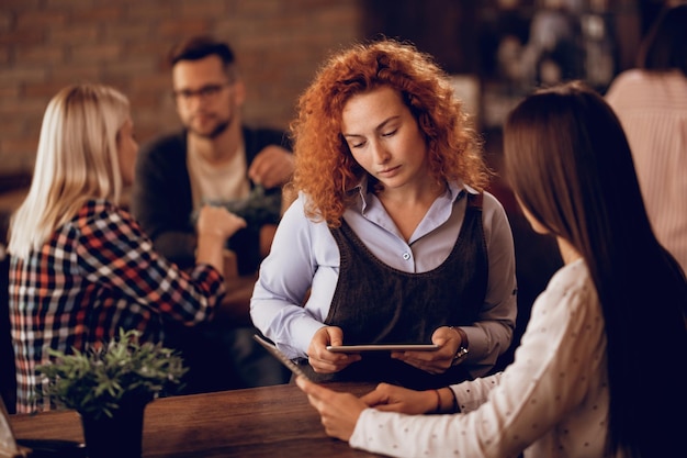 Jeune serveuse aidant une invitée à choisir quelque chose dans le menu tout en prenant une commande dans un pub