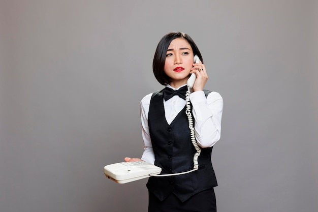 Photo gratuite jeune serveuse asiatique confiante en uniforme parlant au téléphone fixe et détournant les yeux. réceptionniste de femme sérieuse tenant un téléphone rétro et parlant avec le personnel du restaurant