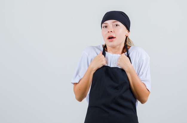 Jeune serveuse en uniforme et tablier gardant les poings sur la poitrine et à la surprise