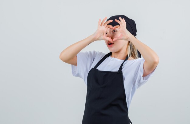 Jeune serveuse en uniforme et tablier montrant le geste du cœur sur les yeux