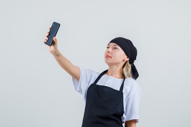 Jeune serveuse en uniforme et tablier prenant selfie sur téléphone mobile