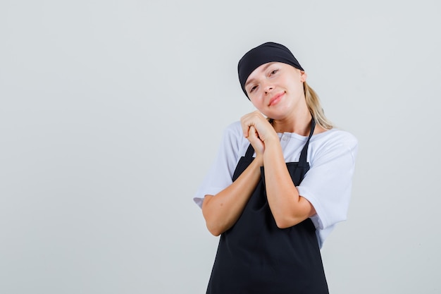 Jeune serveuse en uniforme et tablier s'inclinant la tête sur les mains jointes et à la bien-aimée