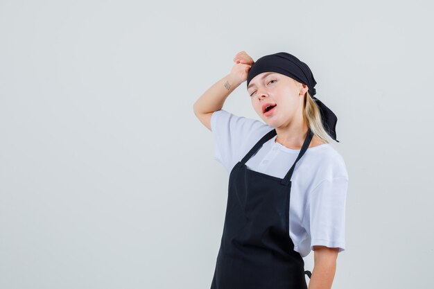 Jeune serveuse en uniforme et tablier se penchant la tête sur la main et clignant des yeux