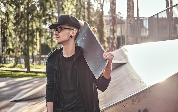 Jeune skateur élégant dans un sweat à capuche noir et une casquette est assis sur la rampe du skatepark en été