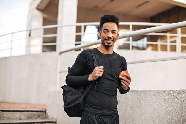 Jeune sportif en t-shirt à manches longues noir tient un sac à dos et une pomme fraîche