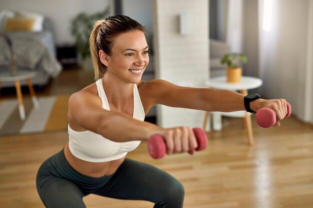 Jeune sportive heureuse faisant des squats tout en faisant de l'exercice avec des poids dans les mains dans le salon