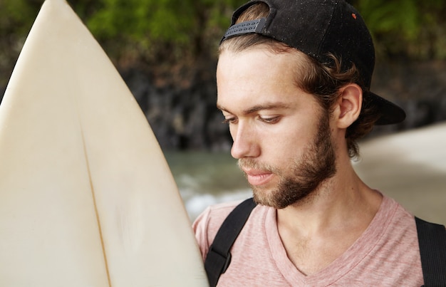 Jeune surfeur pro barbu sérieux et concentré en snapback tenant sa planche de surf, se décidant avant le concours de surf