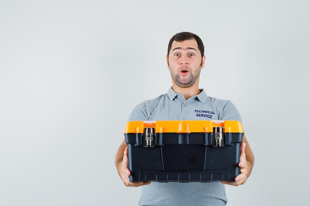 Jeune technicien présentant une boîte à outils en uniforme gris et à la surprise.