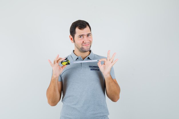 Jeune technicien en ruban à mesurer d'ouverture uniforme gris et à la gaieté.