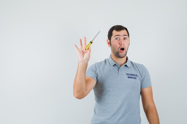 Jeune technicien tenant des tournevis dans une main en uniforme gris et à la surprise.