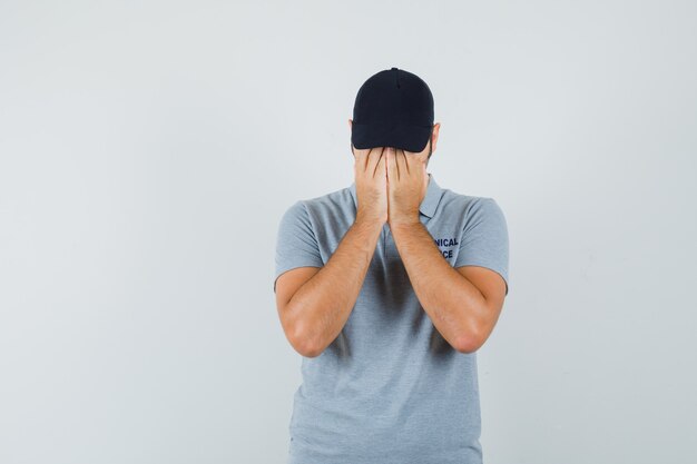 Jeune technicien en uniforme gris couvrant le visage avec les mains et l'air déprimé.