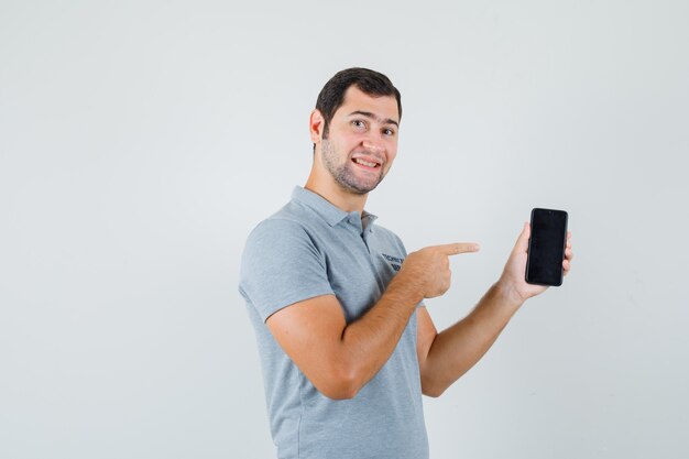 Jeune technicien en uniforme gris pointant sur le téléphone mobile et à la joyeuse vue de face.