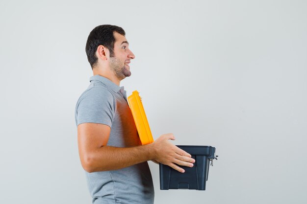 Jeune technicien en uniforme gris portant une boîte à outils ouverte avec ses deux mains et à l'optimisme.